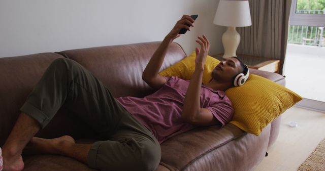A young man in casual clothing lying on a couch with yellow pillows, wearing headphones and using a smartphone. Perfect for themes related to relaxation, technology use, digital lifestyle, and home comfort.