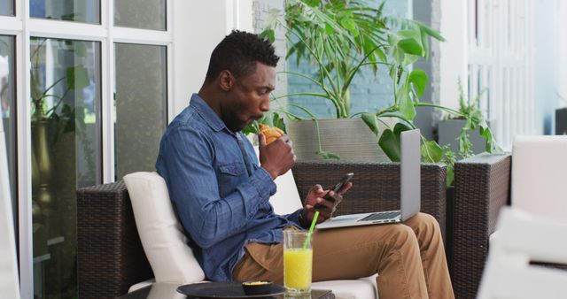 Young man enjoying remote work from a patio, multitasking with his laptop and smartphone while eating a sandwich. The scene includes greenery and orange juice, suggesting a relaxed and productive environment ideal for work-related content, lifestyle blogs, and advertisements for remote work solutions.