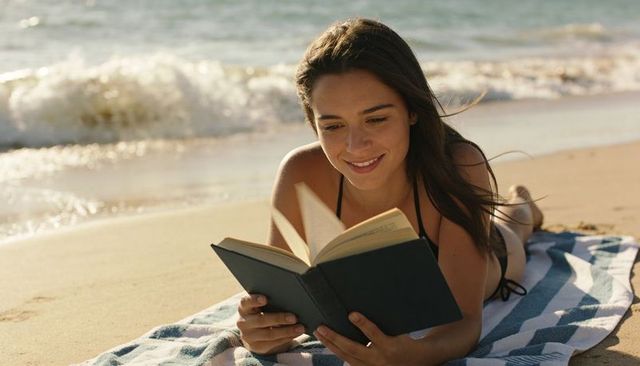 Young woman reading hardcover book on beach towel by sunlit waves summer relaxation