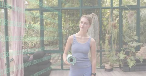 Young woman standing holding rolled yoga mat in sunlit glass greenhouse for wellness