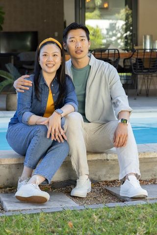 Young diverse couple relaxing by backyard pool wearing smartwatches and white sneakers