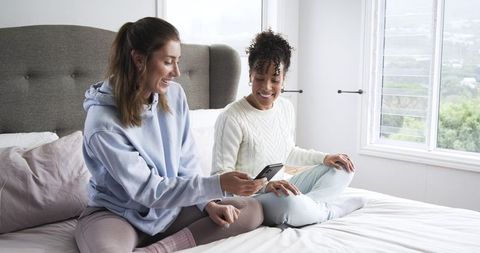 Diverse female friends sitting on bed sharing smartphone and smiling in cozy bedroom