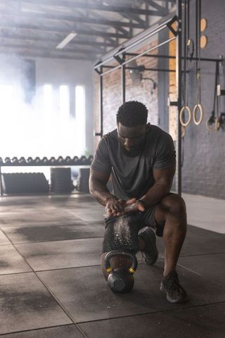 African American Athlete Kneeling Dusting Hands with Chalk over Kettlebell in Industrial Gym