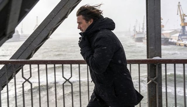 Solitary man bracing wind walking snowy harbor bridge, gripping railing, wearing coat