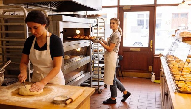 Female baker shaping dough while colleague removes croissants from oven in artisan bakery