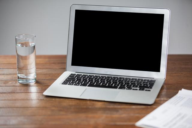 Laptop with a blank screen next to a glass of water placed on a wooden table. Ideal for illustrating office setups, remote work environments, or articles on maintaining hydration while working. Suitable for blog posts, websites about technology, productivity, and health tips for working professionals.