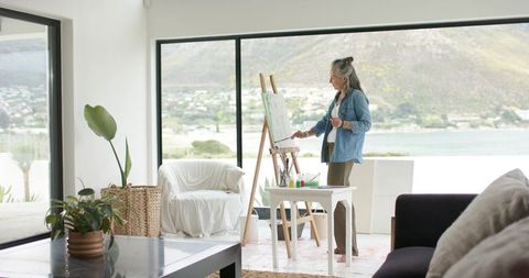 Mature woman painting at easel in sunlit coastal living room, cozy airy artist studio