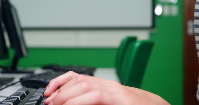 Close-up view of hands typing on a computer keyboard with a green wall in the background. Ideal for use in educational content, technology training materials, presentations on modern learning environments, or work-place training programs.