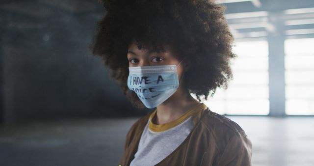 Depicting a woman with afro hair wearing a face mask with the words 'I have a voice.' The background shows a dimly lit urban indoor space. Useful for themes about social awareness, public health, activism, pandemic, and personal expression.