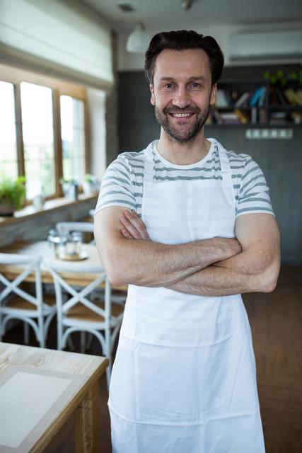 A male bakery owner smiling confidently while standing with crossed arms in his shop. The bright and inviting interior adds warmth to the scene. This image can be used for promoting bakery businesses, articles on entrepreneurship, customer service, or hospitality services.