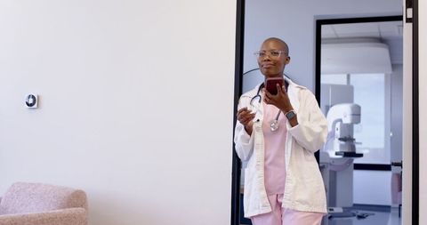 Clinician standing using smartphone at clinic wearing pink scrubs and lab coat for telehealth