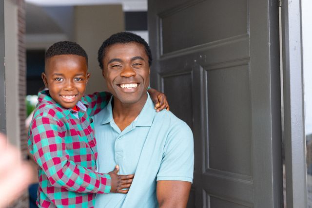 This image depicts a joyful African American father holding his son while standing at the front door, both smiling warmly. Ideal for use in family-oriented advertisements, parenting blogs, and promotional materials highlighting family values, home life, and positive relationships.