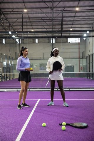 Diverse female athletes practicing padel on purple court holding racket and balls