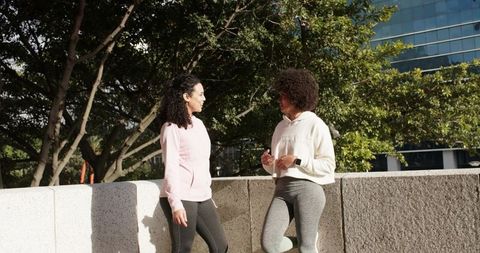 Two Women Chatting in Urban Green Plaza Wearing Hoodies and Leggings on Sunny Morning