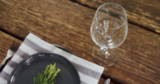 An empty wine glass sits beside a dark plate with a sprig of greenery on a wooden table, with copy space. The setting suggests a rustic dining experience or the preparation for a meal.
