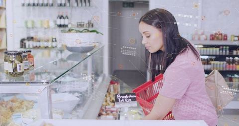 Young woman selecting fresh pastries at deli counter with red basket in bright market
