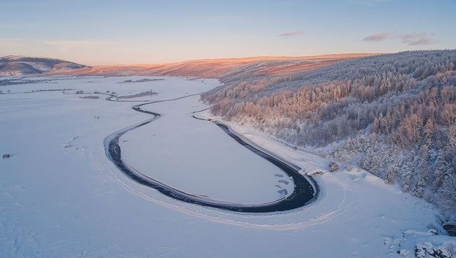 Sunrise lighting frozen meandering river loop along snow-covered forest aerial winter vista