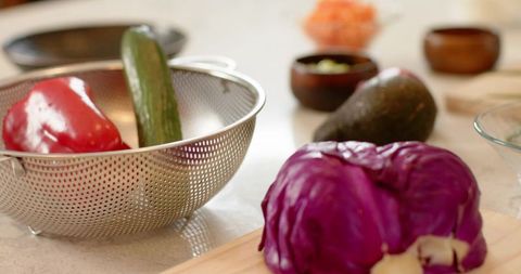 Purple cabbage sitting on wooden cutting board with colander of red pepper and cucumber