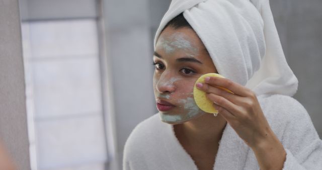 Woman applying face mask in front of bathroom mirror. She is wearing a white towel turban and robe, suggesting a moment of self-care and pampering. This image is suitable for content related to skincare routines, beauty tips, wellness, and self-care promotions.