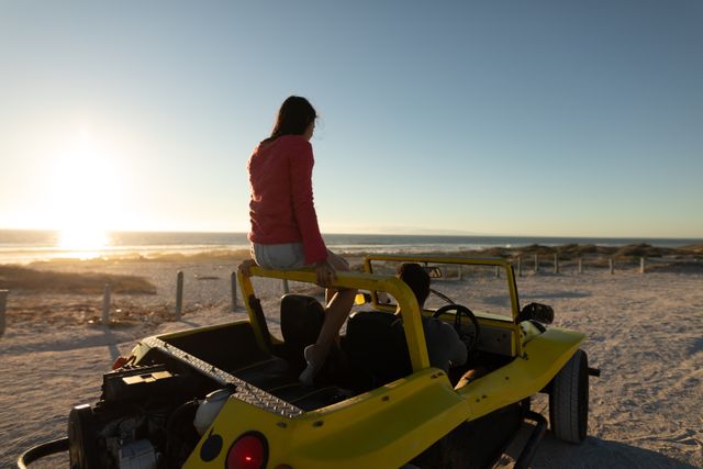 Happy caucasian couple driving in beach buggy by the sea at sunset, woman sitting on roll bar. beach stop off on romantic summer holiday road trip.