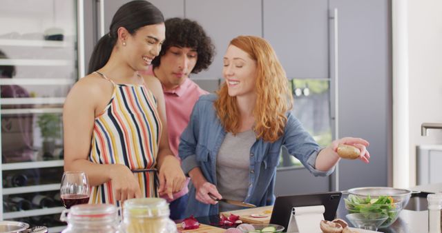 Group of friends cooking together and enjoying meal preparation in a modern kitchen. They are chopping vegetables and using a tablet for a recipe. Ideal for concepts like social interaction, teamwork in cooking, culinary hobbies, and friendship.