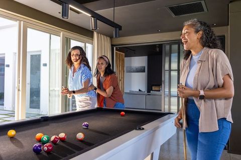 Multicultural women laughing and playing pool while holding cues in modern home game room daylight