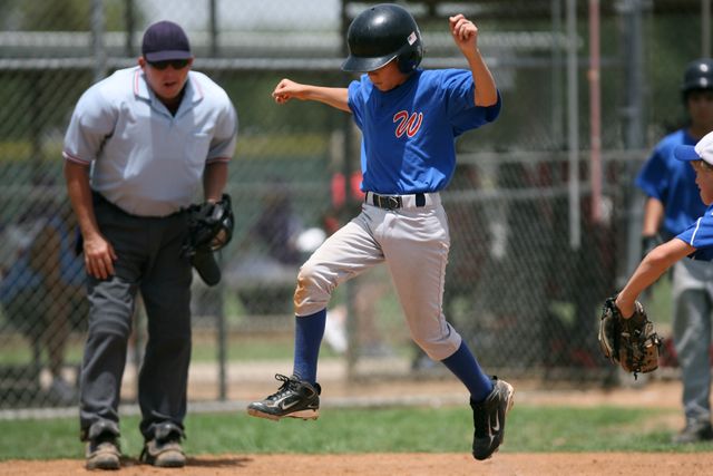 Youth baseball player jumping in excitement during an outdoor game. Umpire is observing the action, with a teammate beside him. Perfect for depicting action, sportsmanship, team activities, and youth involvement in sports. Useful for promoting summer sports camps, youth sports leagues, or athletic events.