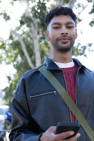 Young man standing on sidewalk holding smartphone, wearing leather jacket and red sweater