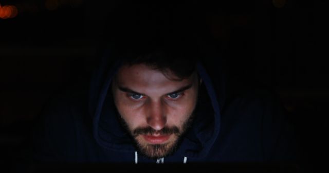 Young man in a dark room, wearing a hoodie, intensely focused on a computer screen. Suitable for use in articles or advertisements related to cybersecurity, programming, online gaming, late-night work, or technology addiction.
