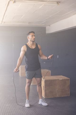 Fit athletic man skipping rope in industrial gym wearing smartwatch and performance shorts