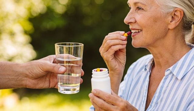 Senior woman taking capsule outdoors while caregiver offers glass of water in sunny garden