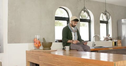 Indian man using tablet on kitchen island with beanie and coffee, minimalist kitchen