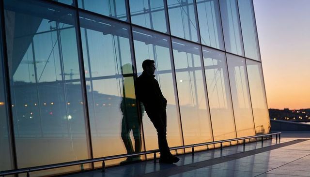 Man leaning against glass atrium reflecting city lights at twilight