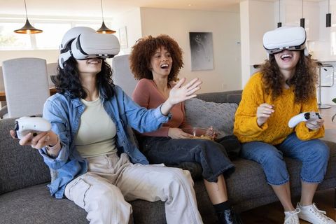 Diverse friends wearing VR headsets laughing and playing multiplayer games on sofa at home