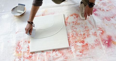 Indian artist kneeling spreading gesso with trowel on canvas over paint-stained drop cloth