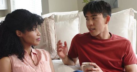 Multiracial couple talking on sofa sharing coffee and honest conversation in sunlit living room