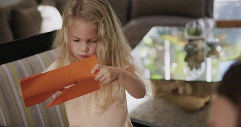 Young girl examining orange paper craft while sitting on striped cushion in living room