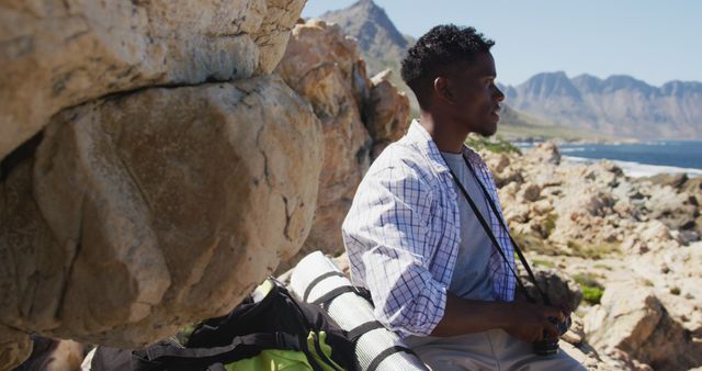 Young black man hiking and enjoying beautiful scenery with mountains and sea in background. Ideal for promoting outdoor activities, adventure tourism, nature exploration, and leisure. Perfect for use in travel blogs, tourism brochures, and advertisements targeting outdoor enthusiasts.