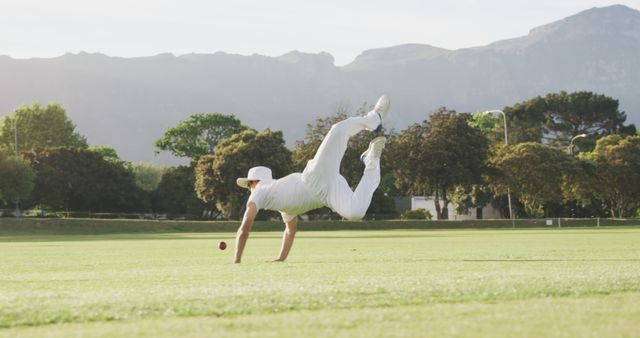 Cricketer dressed in white uniform is diving to catch the ball on a lush green cricket field, with a scenic mountain backdrop. Suitable for use in sports-related content, athletic training articles, motivational posters, outdoor activities promotions, and cricket event advertisements.