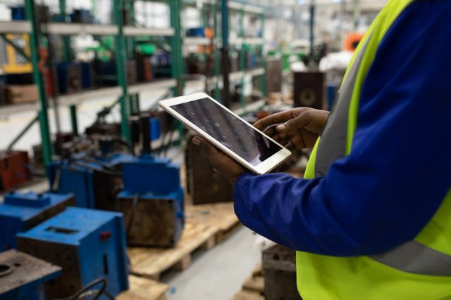 Side view mid section of a biracial male worker working in a busy factory warehouse wearing a hi vis vest and blue overalls, standing and using a tablet computer