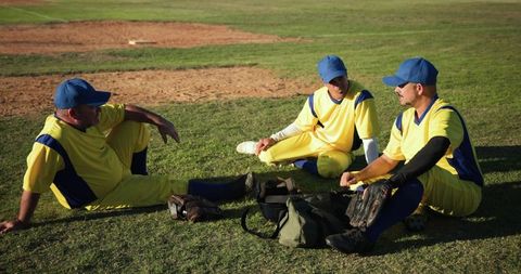 Veteran baseball teammates relaxing and chatting on grass in yellow-blue uniforms after game