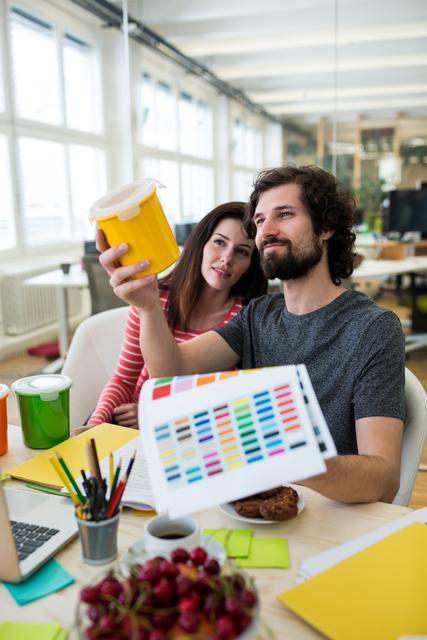 Male and female graphic designers holding plastic container and color chart in office