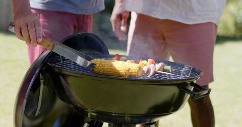 Family cooking corn and vegetable skewers on charcoal grill outdoors