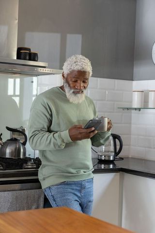 Senior african american man checking smartphone while holding mug in modern kitchen