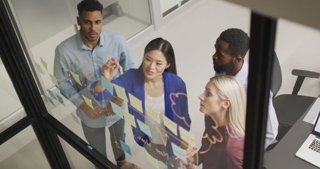 Diverse team of professionals collaborating on a project using sticky notes on a glass wall in a modern office. This image is perfect for websites and articles related to teamwork, project management, corporate culture, and innovative business strategies.
