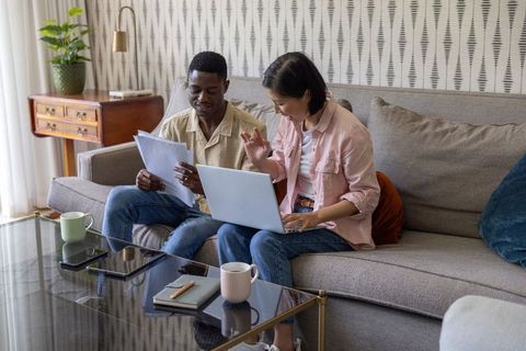 Mixed-race couple managing household finances with laptop and paperwork on sofa