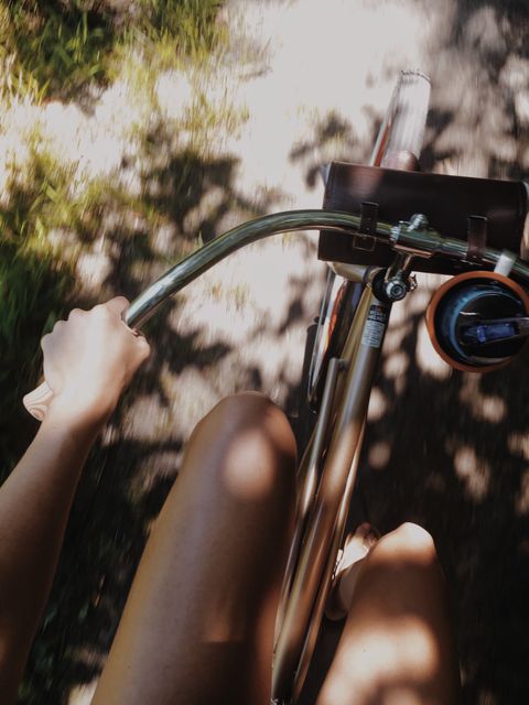 Person seen from a first-person perspective riding a vintage bike on a sunlit path. Their hands are holding the handlebars, capturing a sense of freedom and relaxation. This depiction of a leisure activity in nature highlights the joy of cycling and can be used in content related to healthy lifestyles, outdoor activities, summer adventures, and sustainable transportation.