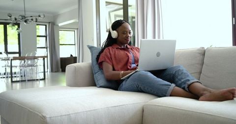 African american woman reclining on sofa using laptop and headphones in modern living room