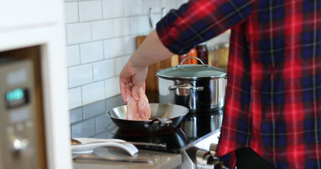 Person placing chicken breast in frying pan on stovetop in modern kitchen. Stainless steel pot with lid is visible on stove. Wearing red plaid shirt. Useful for cooking tutorials, blogs on home-made recipes, kitchen appliance advertisements, and articles on domestic culinary practices.