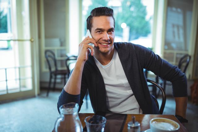 Young man sitting in a café, smiling while talking on his mobile phone. Ideal for use in advertisements for mobile phone services, lifestyle blogs, or articles about communication and technology.